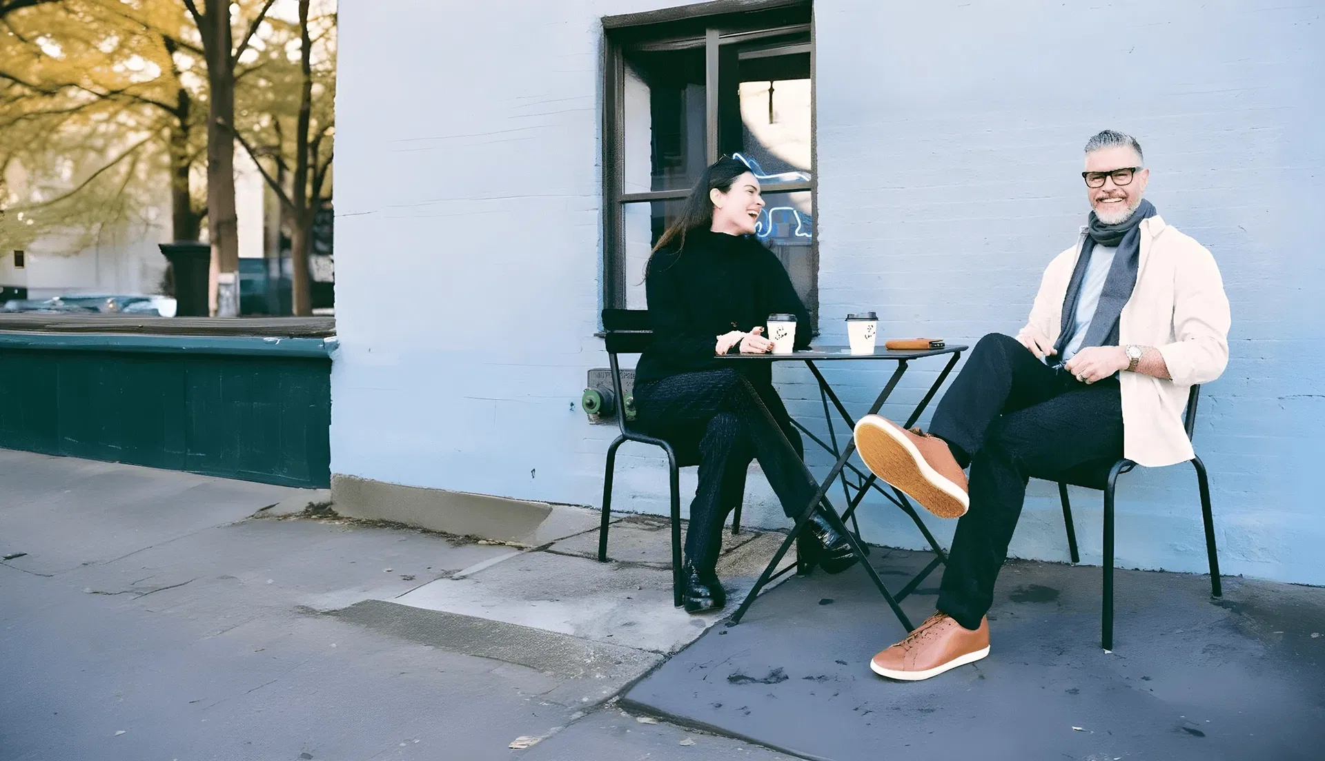 Two people sit at an outdoor table, smiling and drinking coffee, with a blue wall behind them.