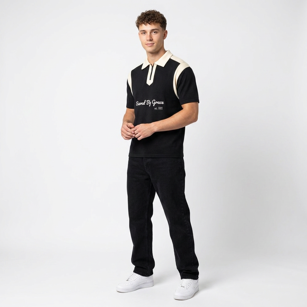 A young man wearing a black and white polo shirt with text, black pants, and white sneakers standing against a plain background