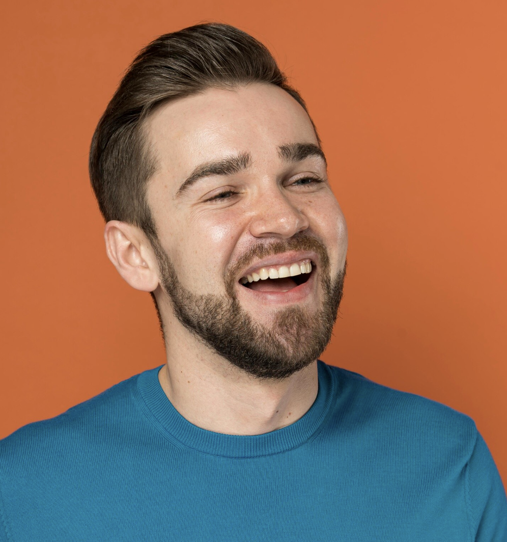 Smiling young man with beard wearing a turquoise shirt against an orange background.
