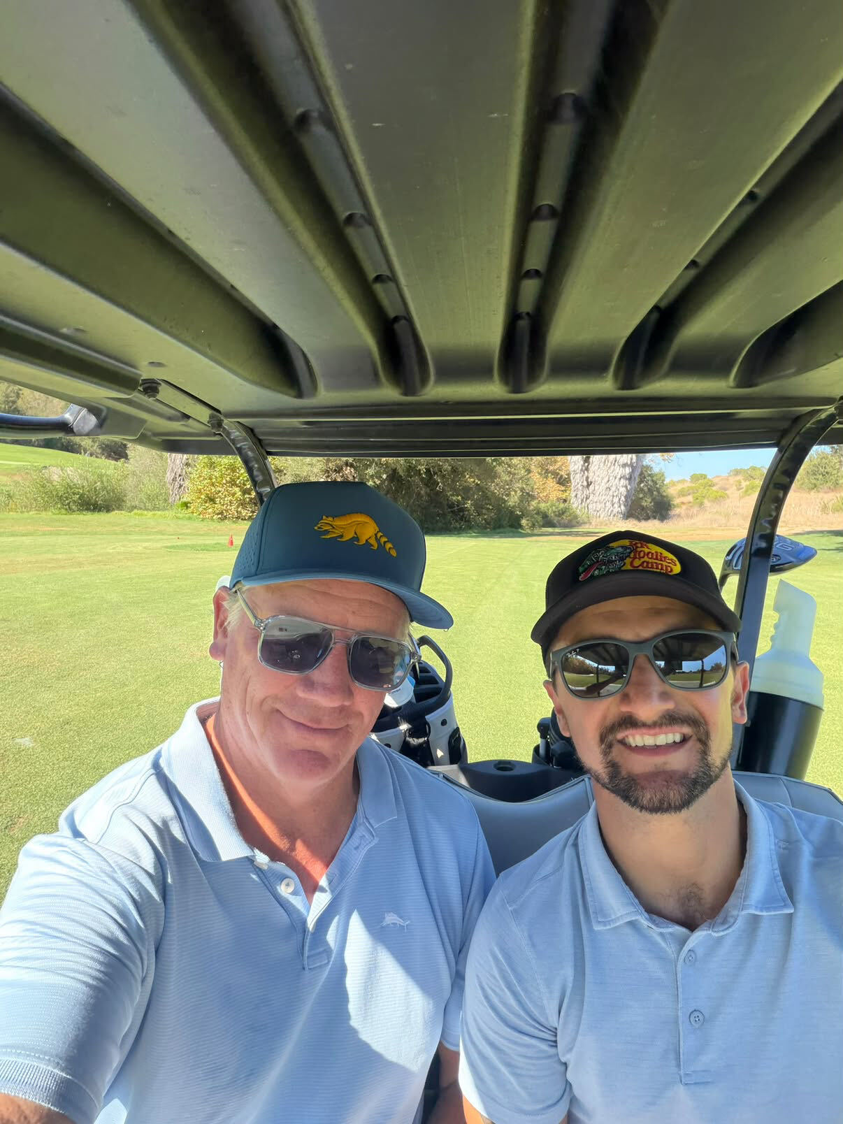 Two men in caps and light blue polos taking a selfie in a golf cart on a sunny course, wearing St Barts Azure Polarised sunglasses