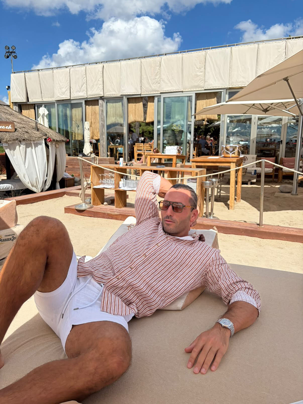 Man in a striped linen shirt and white shorts reclining on a sunbed at a beach club, wearing St Barts Safari Polarised sunglasses with brown lenses