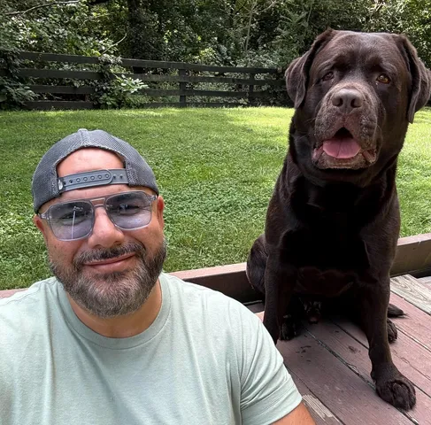 Bearded man in a backwards cap and sage t-shirt taking a backyard selfie with his chocolate Labrador, wearing St Barts Azure sunglasses