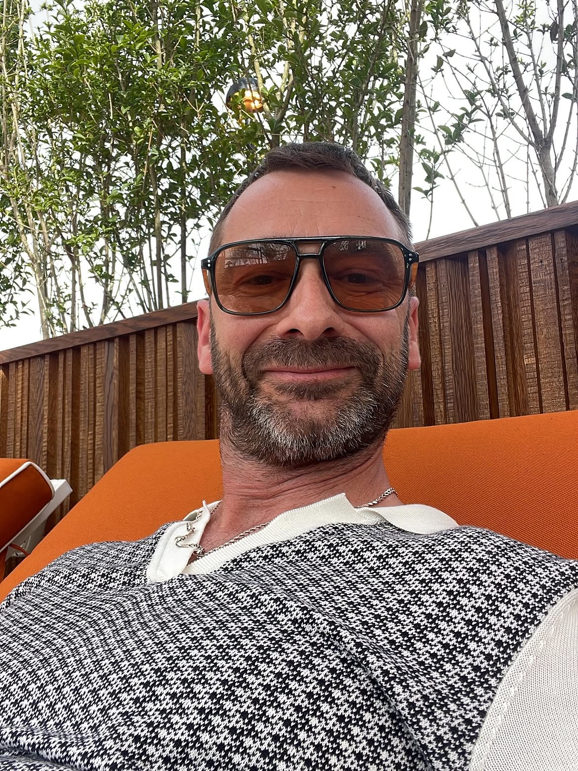 Smiling bearded man in a houndstooth knit relaxing on an orange rooftop lounger, wearing St Barts Safari sunglasses with amber lenses