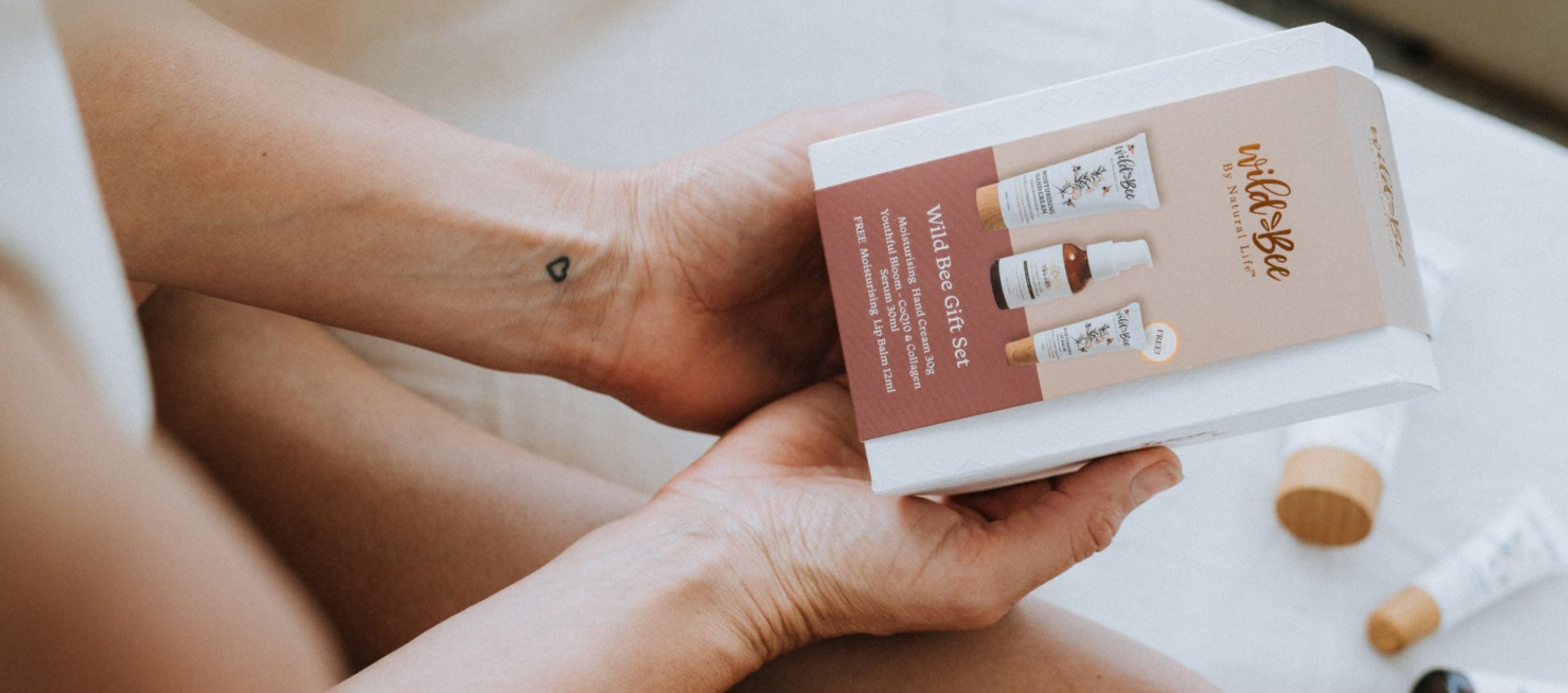 Woman sitting on a bed holding a Wild Bee gift set box, with hand cream and lip balm beside her