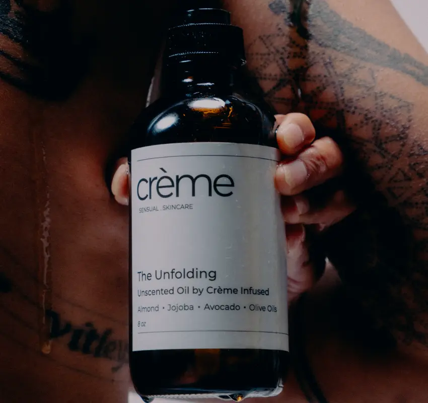 Close-up of "The Unfolding Oil" bottle being held against a tattooed body, highlighting the brand’s focus on authentic personal care rituals for real bodies.