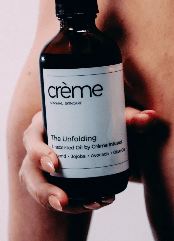 A close-up of a hand holding a bottle of "The Unfolding" unscented oil against bare skin, illustrating the brand's mission to inspire women to come home to their bodies through touch.