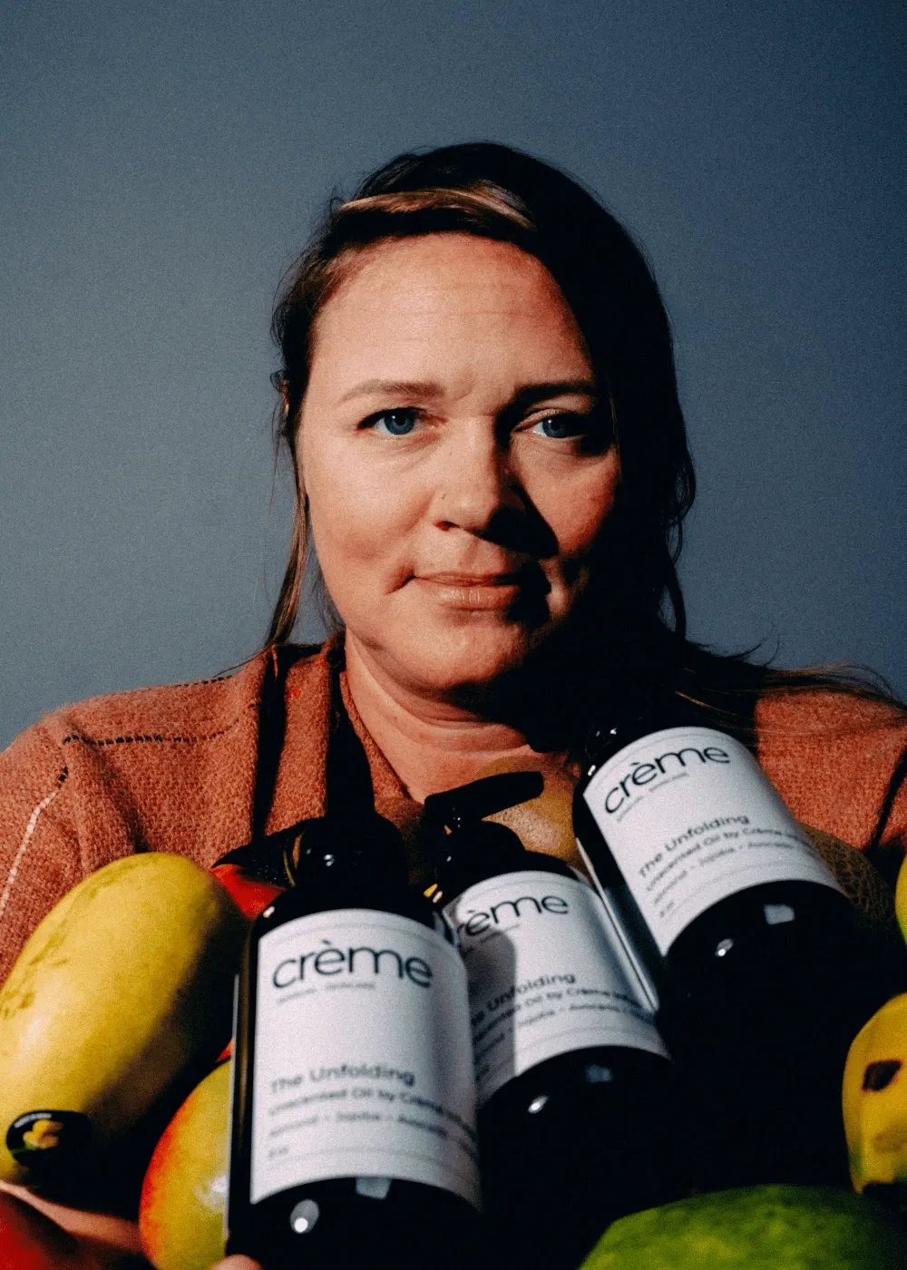 A warm portrait of founder Samantha holding bottles of "The Unfolding Oil" and fresh fruit, celebrating the vibrant "Juicy Middle" of life and the evolving feminine journey.