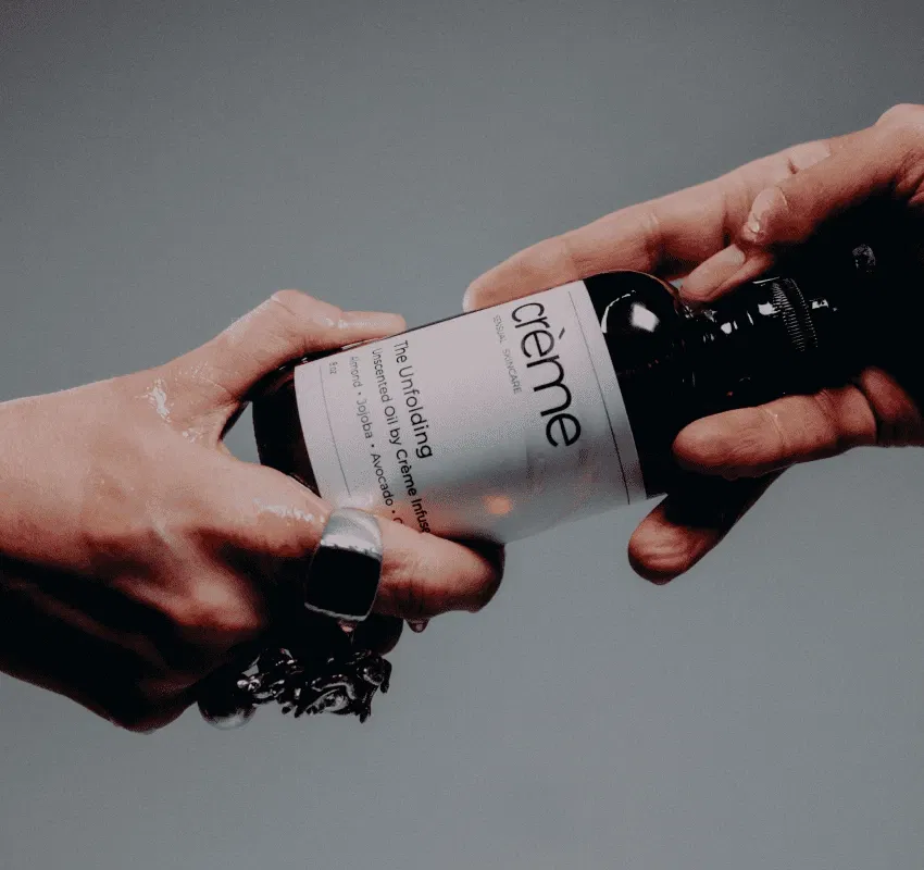 Two sets of hands passing a bottle of "The Unfolding Oil," symbolizing a shared sacred ritual that restores presence and fosters connection.