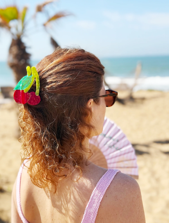 By Vivi Haarklammer Kirsche in roten und grünen Farben als dekorativer Haarclip im lockigen Haar einer Frau am Strand mit Meerblick und sommerlicher Atmosphäre.