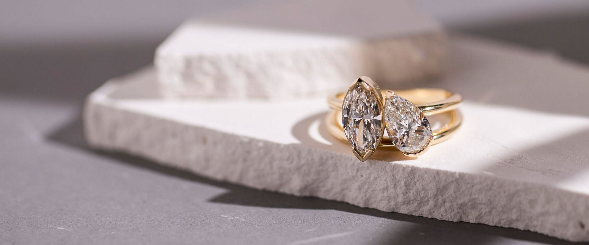 Marquise and pear-cut diamond engagement rings in yellow gold, displayed on stacked white stone slabs with soft natural shadows.