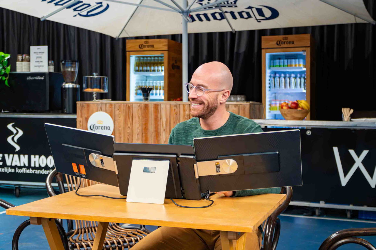 Smiling man using triple-portable monitor setup at a table under a Corona umbrella in a cafe.