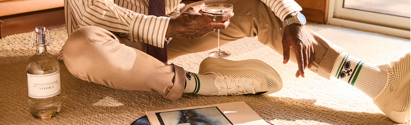 A man relaxing on a woven rug in warm sunlight with a cocktail, a bottle of Stateside Vodka beside him