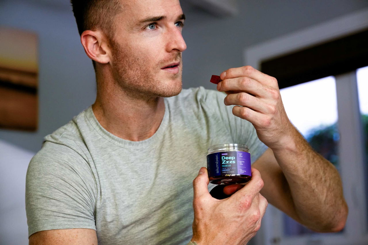 Man in gray shirt holding a gummy and jar labeled &quot;Deep Zzzs&quot; indoors.