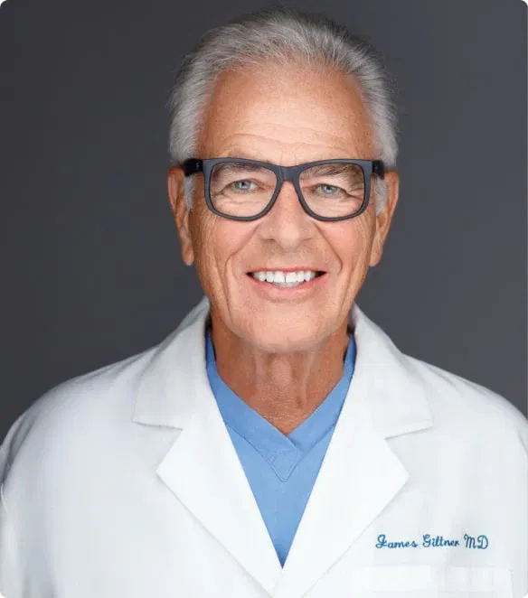 Elderly man with glasses, smiling, wearing a white doctor's coat and blue shirt.