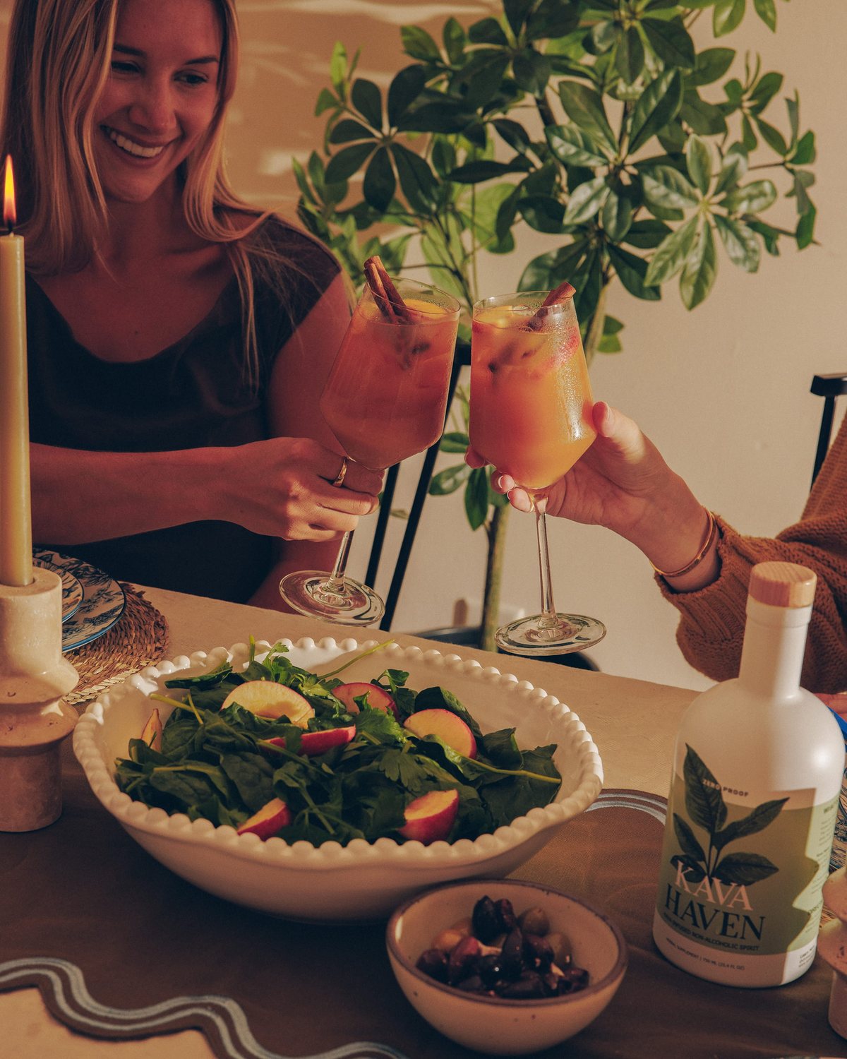 Two friends toasting citrus Kava Haven mocktails over a dinner table set with salad, olives and a bottle of Kava Haven