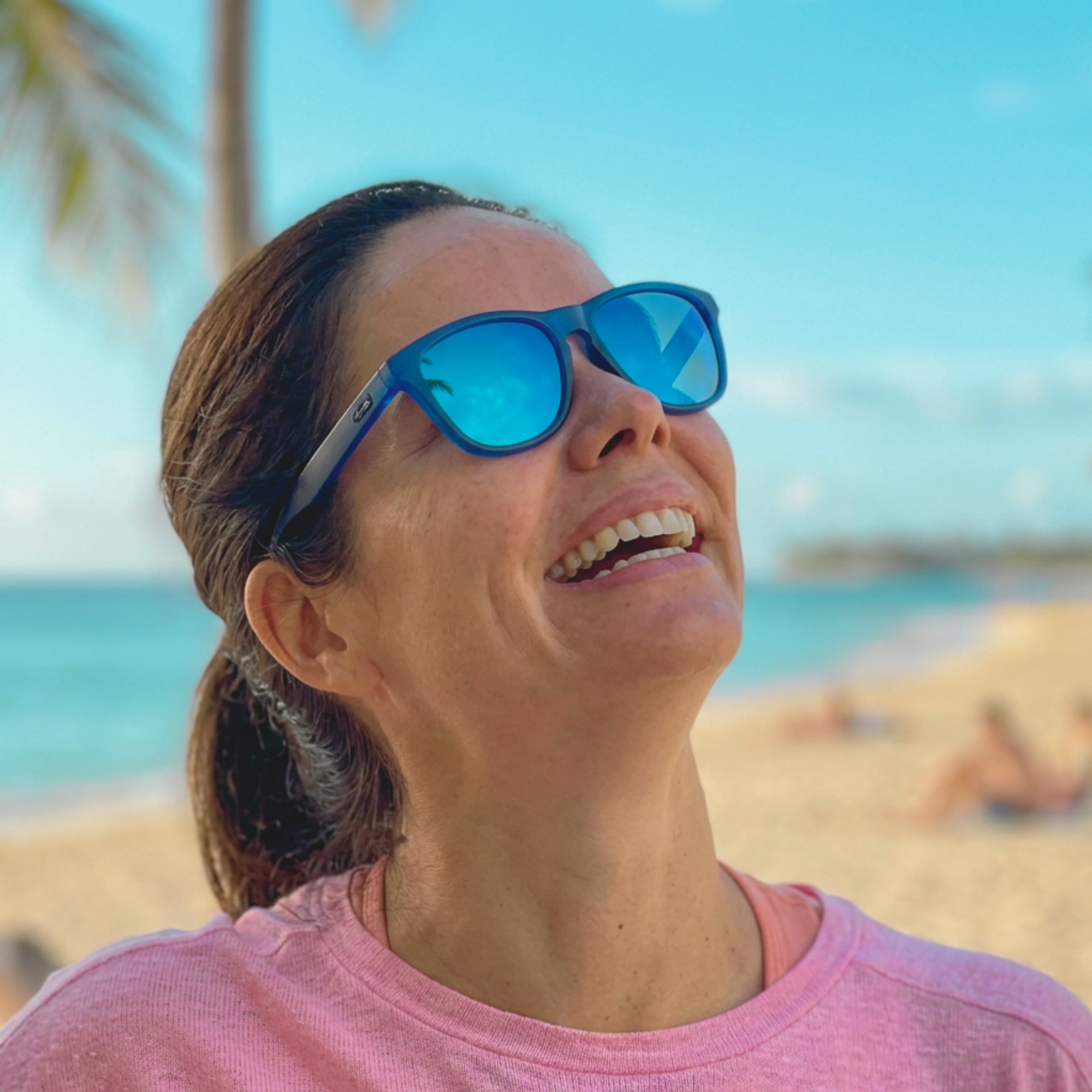 Customer laughing on a tropical beach wearing blue-framed H2OAthletics sunglasses with blue mirror lenses