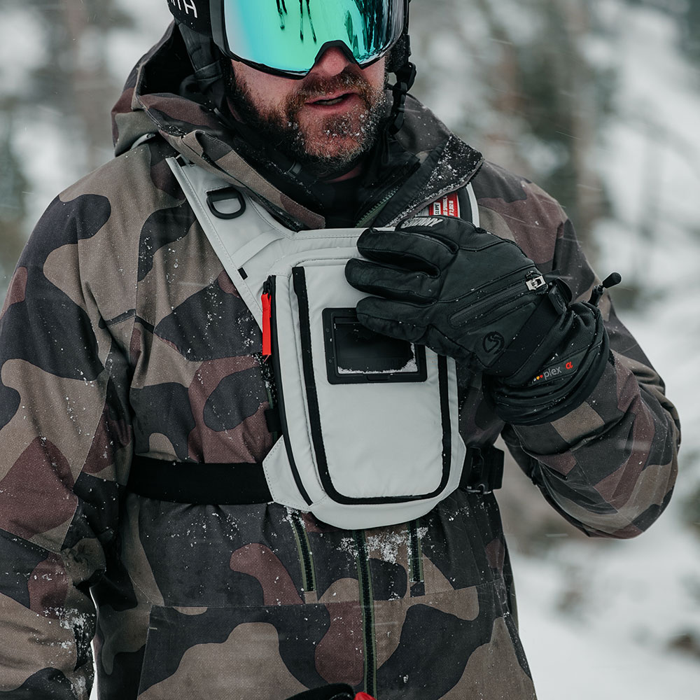 Bearded rider in goggles and a camo snow jacket adjusting the strap of a snow-dusted grey West Slope chest pack with a front phone window in falling snow