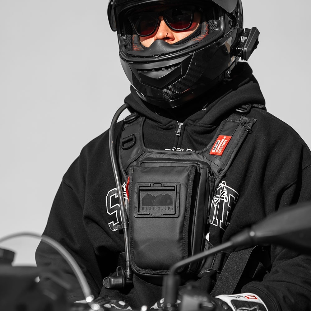 Motorcycle rider in a black full-face helmet and hoodie wearing a black West Slope chest pack with a hydration tube over the shoulder