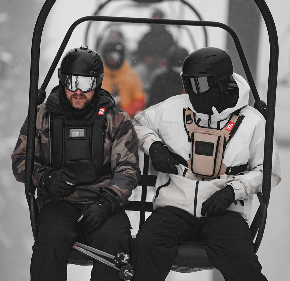 Two riders in helmets and goggles on a snowy chairlift, one wearing a black West Slope chest pack over a camo jacket and the other a tan chest pack over a white shell