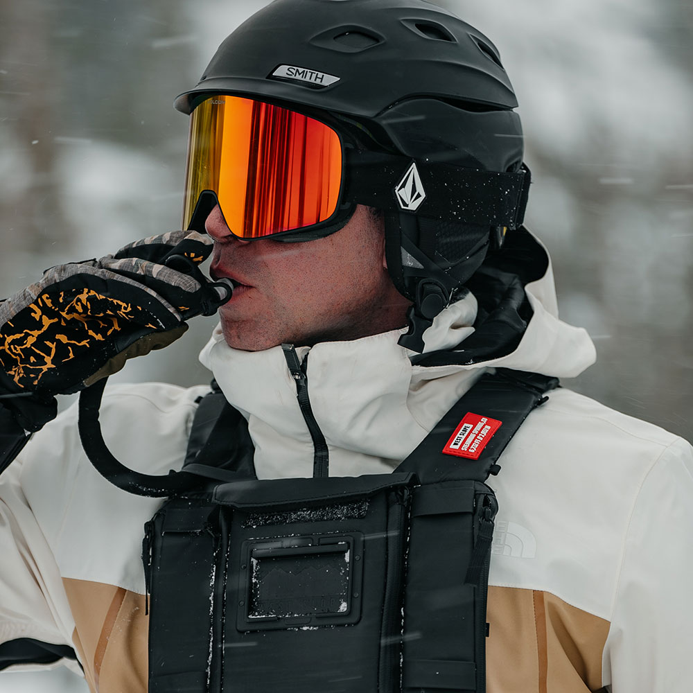 Skier in a black helmet and orange mirrored goggles sipping from the hydration tube of a black West Slope chest pack worn over a white and tan shell in falling snow