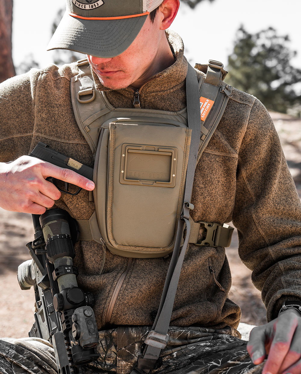Hunter in a brown fleece and camo pants wearing a West Slope chest pack in Coyote Brown, drawing a pistol from the side holster with a scoped rifle at his side