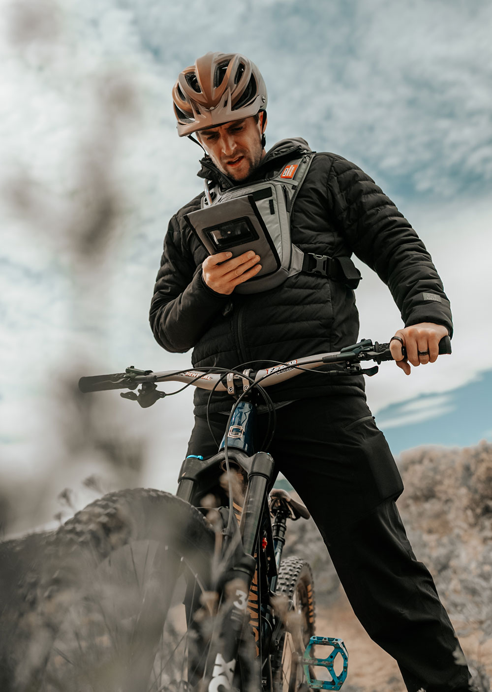 Mountain biker in a bronze helmet and black puffer jacket straddling his trail bike, checking his phone in the window of a grey West Slope chest pack