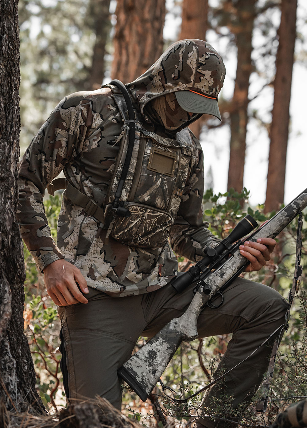 Hunter in camo with a scoped rifle stepping through pine timber, wearing a West Slope chest pack in RealTree Camo with a hydration tube over the shoulder