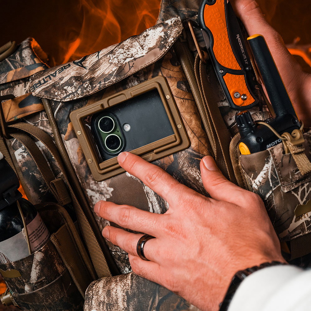 Close-up of a hand on a West Slope chest pack in RealTree Camo, a phone visible through the coyote-brown front window, with a knife and radio clipped to the side pockets