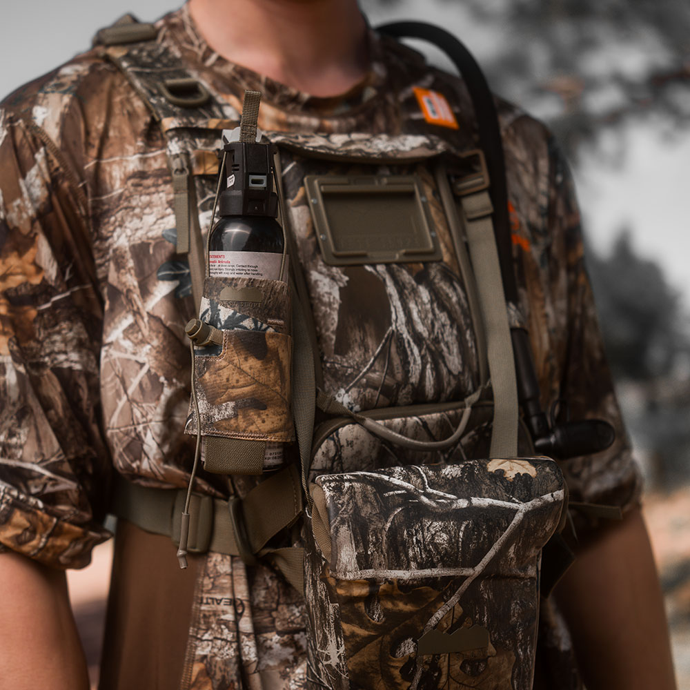 Hunter in RealTree camo wearing a West Slope chest pack in RealTree Camo, with bear spray holstered on the shoulder strap and a hydration tube running over the shoulder