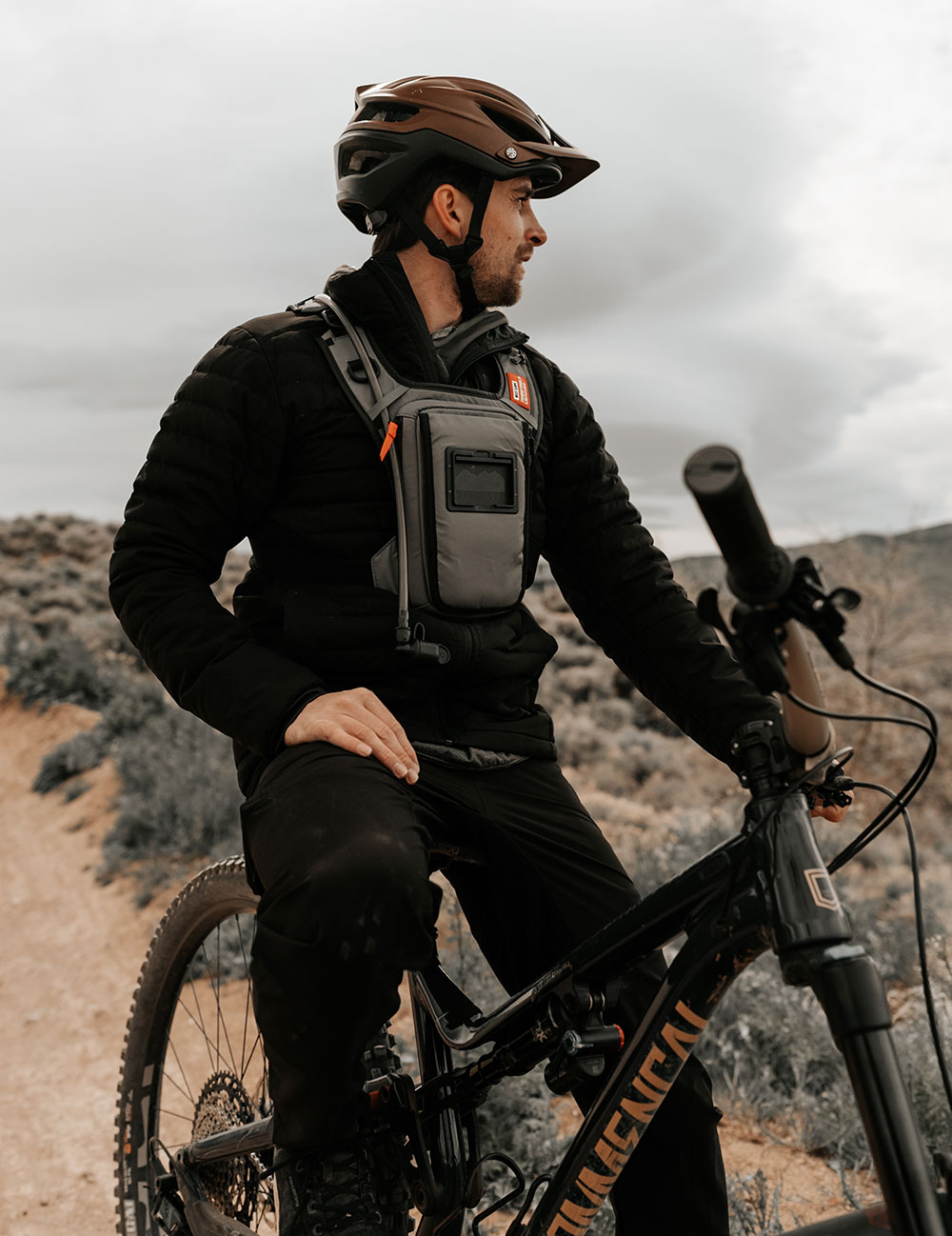 Mountain biker in a brown helmet and black jacket resting on his bike on a desert trail, wearing a grey West Slope chest pack with a phone window