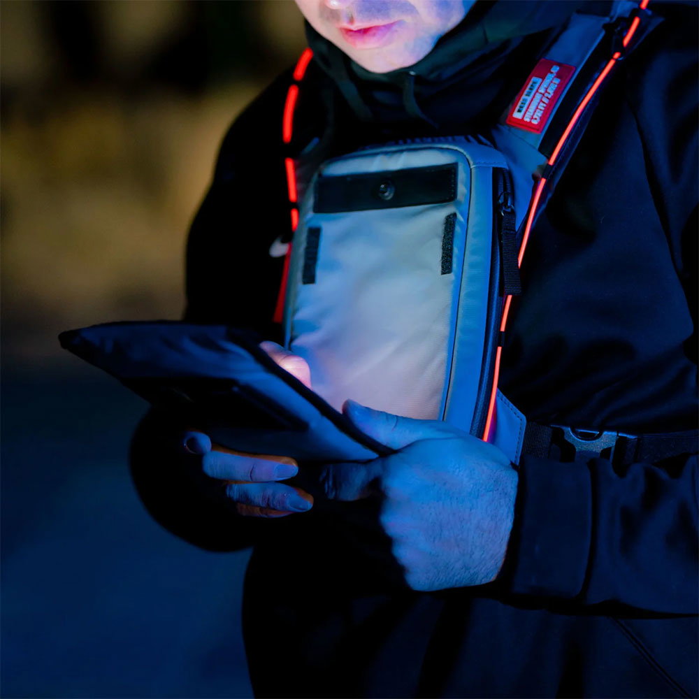 Rider at night opening the front phone window of a grey West Slope chest pack, the Night Kit glowing red along the straps