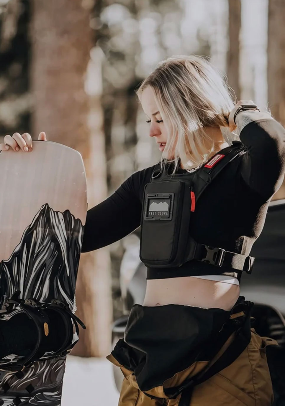 Snowboarder in a black base layer and tan snow pants holding her board among snowy pines, wearing a black West Slope slim chest pack with a front phone window