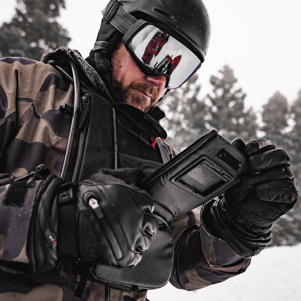 Skier in a black helmet, mirrored goggles and leather gloves flipping open the phone window of a black West Slope chest pack in falling snow