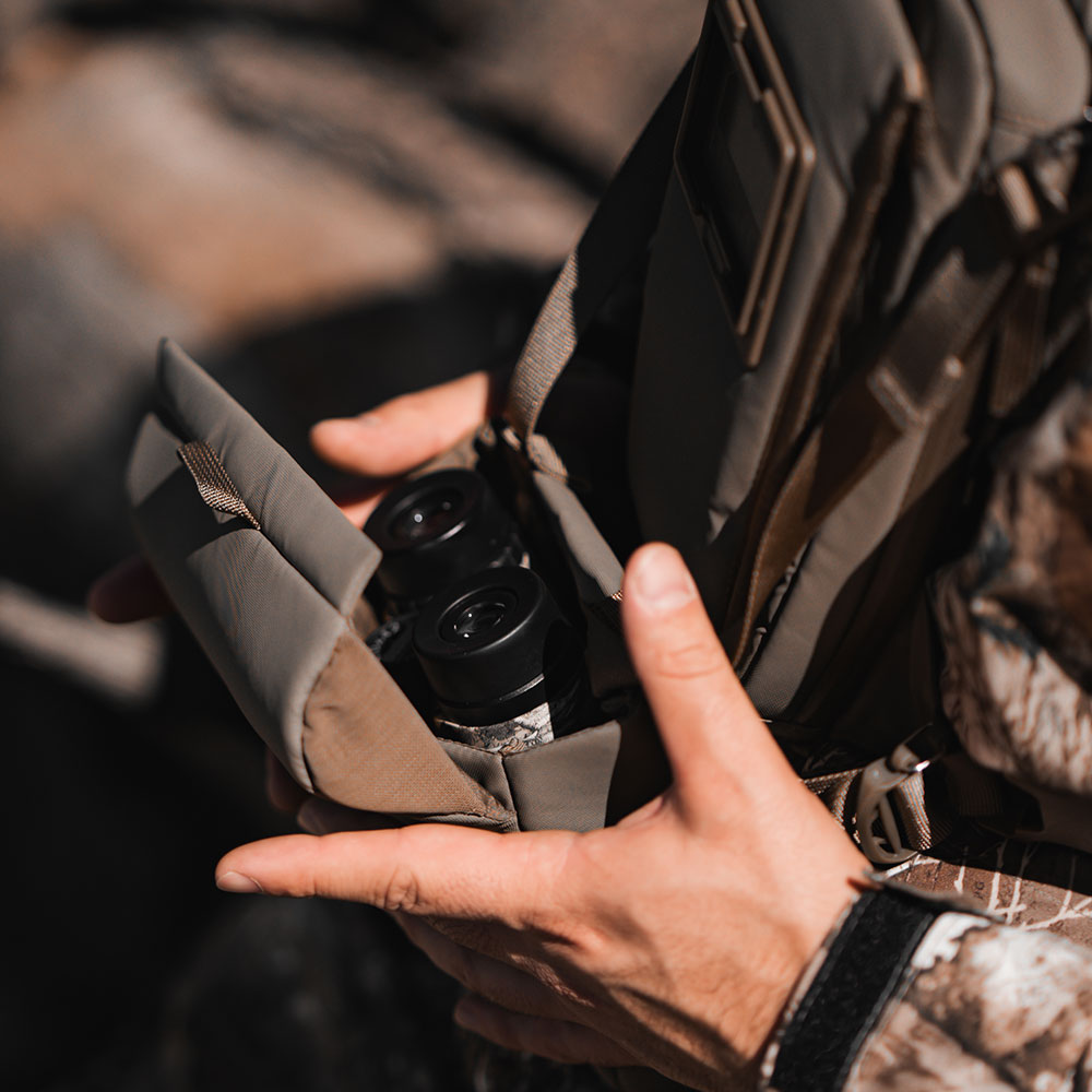 Hunter in camo opening the padded Bino Kit pouch on a West Slope chest pack to reveal a pair of binoculars ready to grab