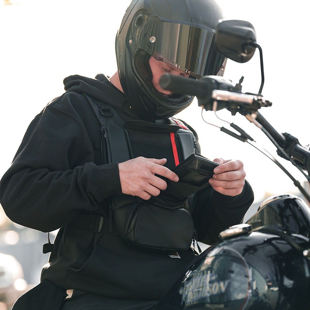Motorcycle rider in a black helmet checking his phone in the front window of a black West Slope chest pack