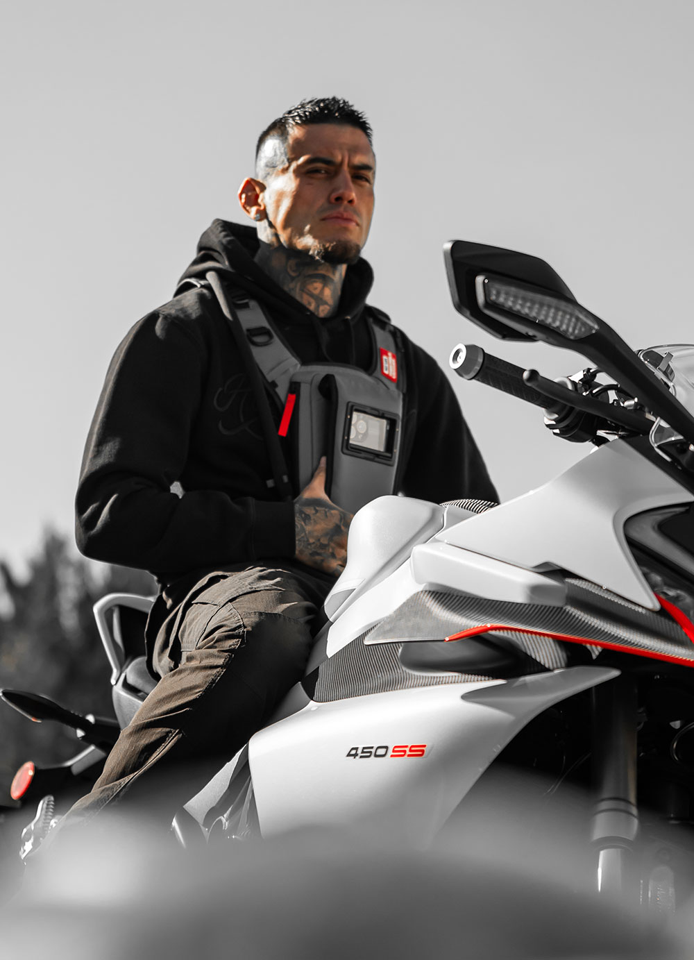 Motorcycle rider in a black hoodie seated on a white sport bike, wearing a grey West Slope slim chest pack with a phone in the front window