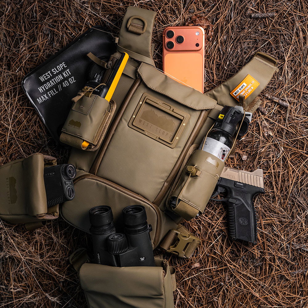 Coyote Brown West Slope chest pack laid out on pine needles with a hydration kit, phone, radio, bear spray, pistol, rangefinder and binoculars stowed in its pockets