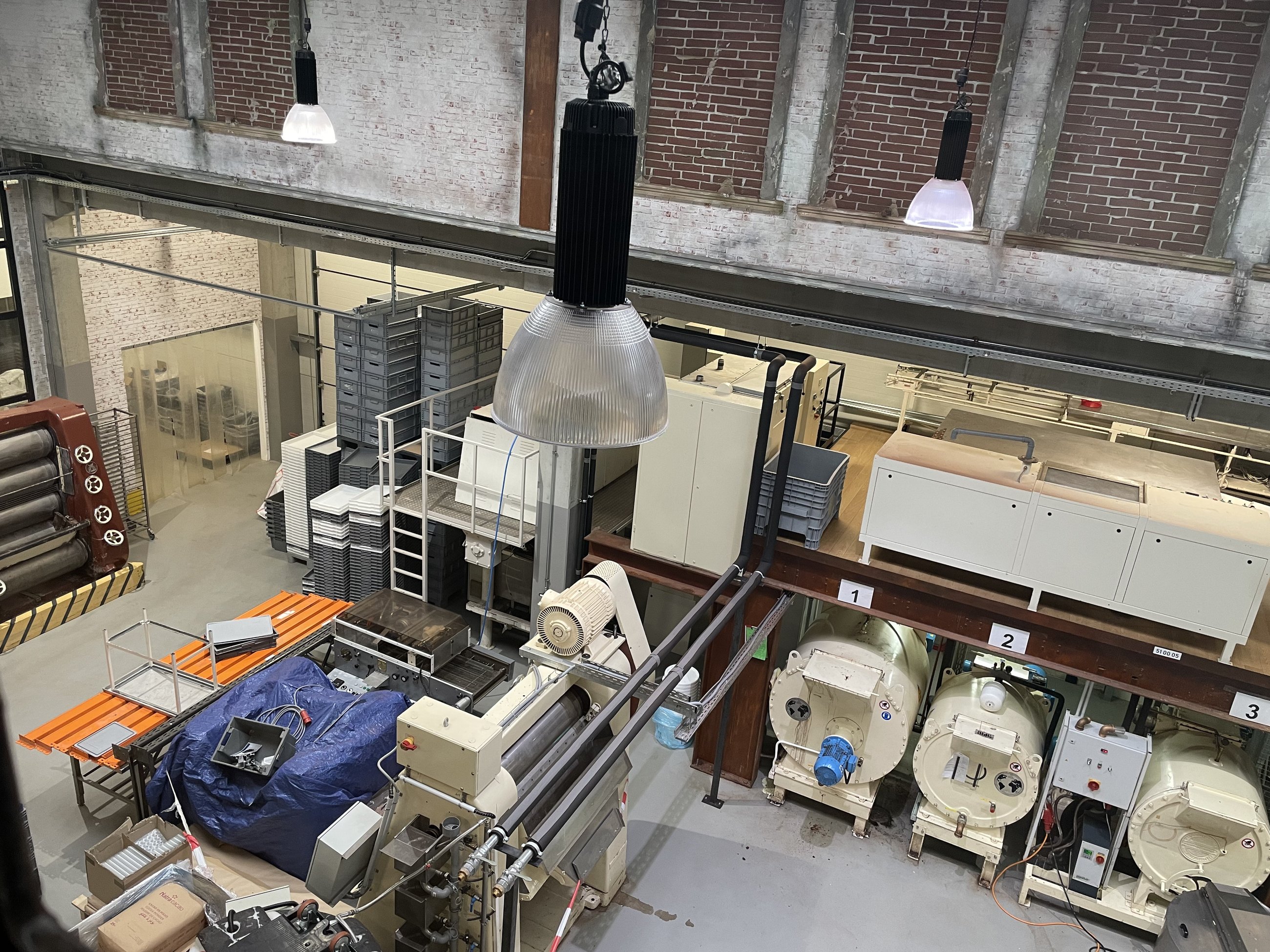 Produktionshalle mit weiß gekalkten Ziegelwänden von oben: unter einer Stahlempore drei cremefarbene, liegende Trommelmaschinen mit den Nummern 1, 2 und 3, vorne eine große Walzenmaschine mit Rohrleitungen