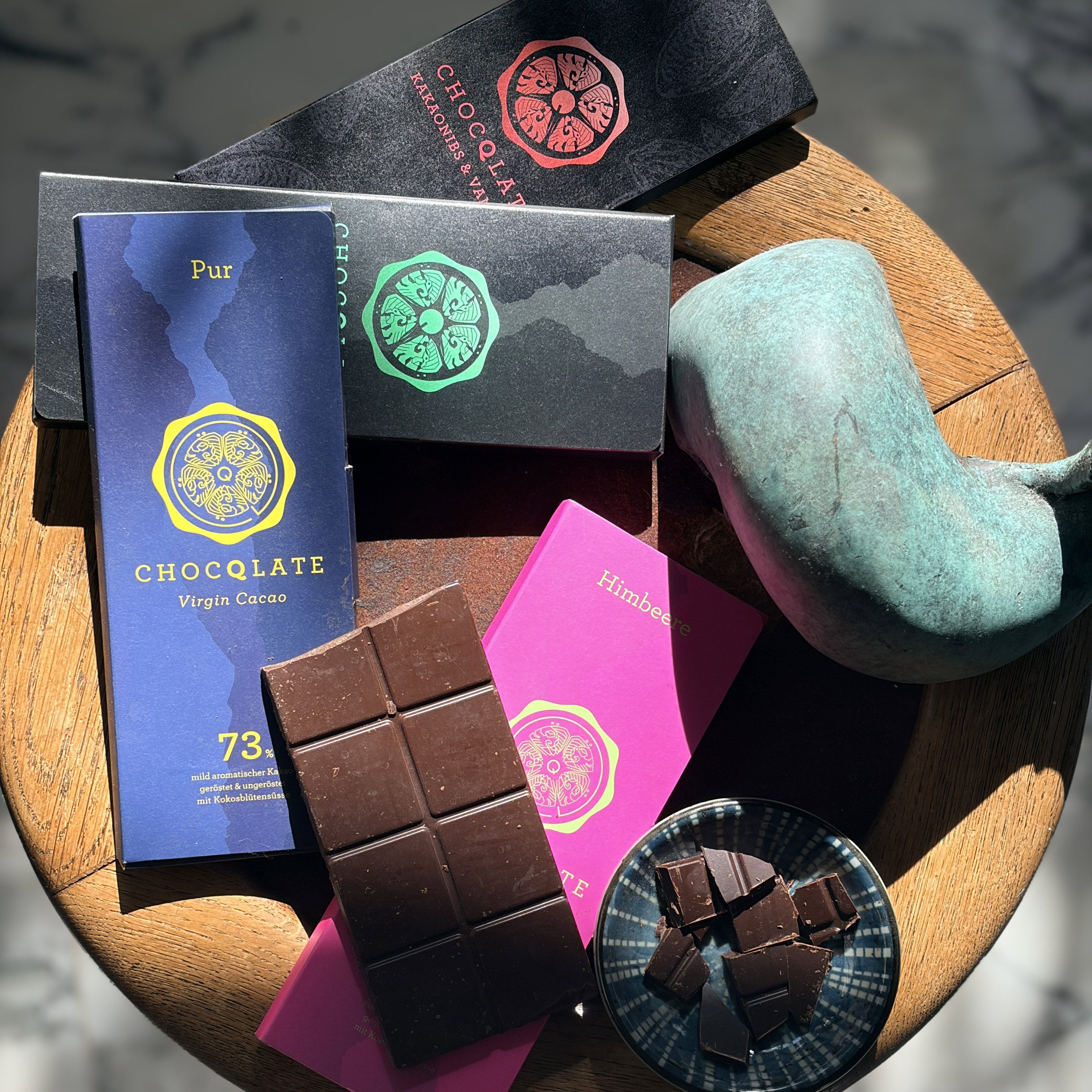 CHOCQLATE Probierpaket – Tafeln Pur 73 %, Himbeere und Kakaonibs & Vanille mit offener Tafel auf einem Holzhocker