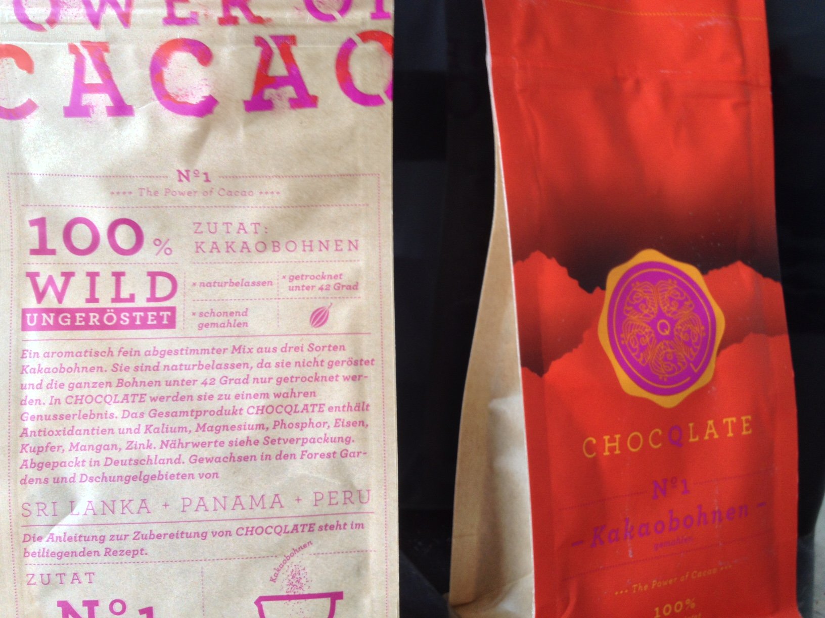 Zwei historische CHOCQLATE-Beutel: links die Rückseite aus Kraftpapier mit der Aufschrift „N°1 · The Power of Cacao · 100 % wild ungeröstet“, rechts die rote Vorderseite mit goldenem Siegel und „N°1 Kakaobohnen gemahlen“