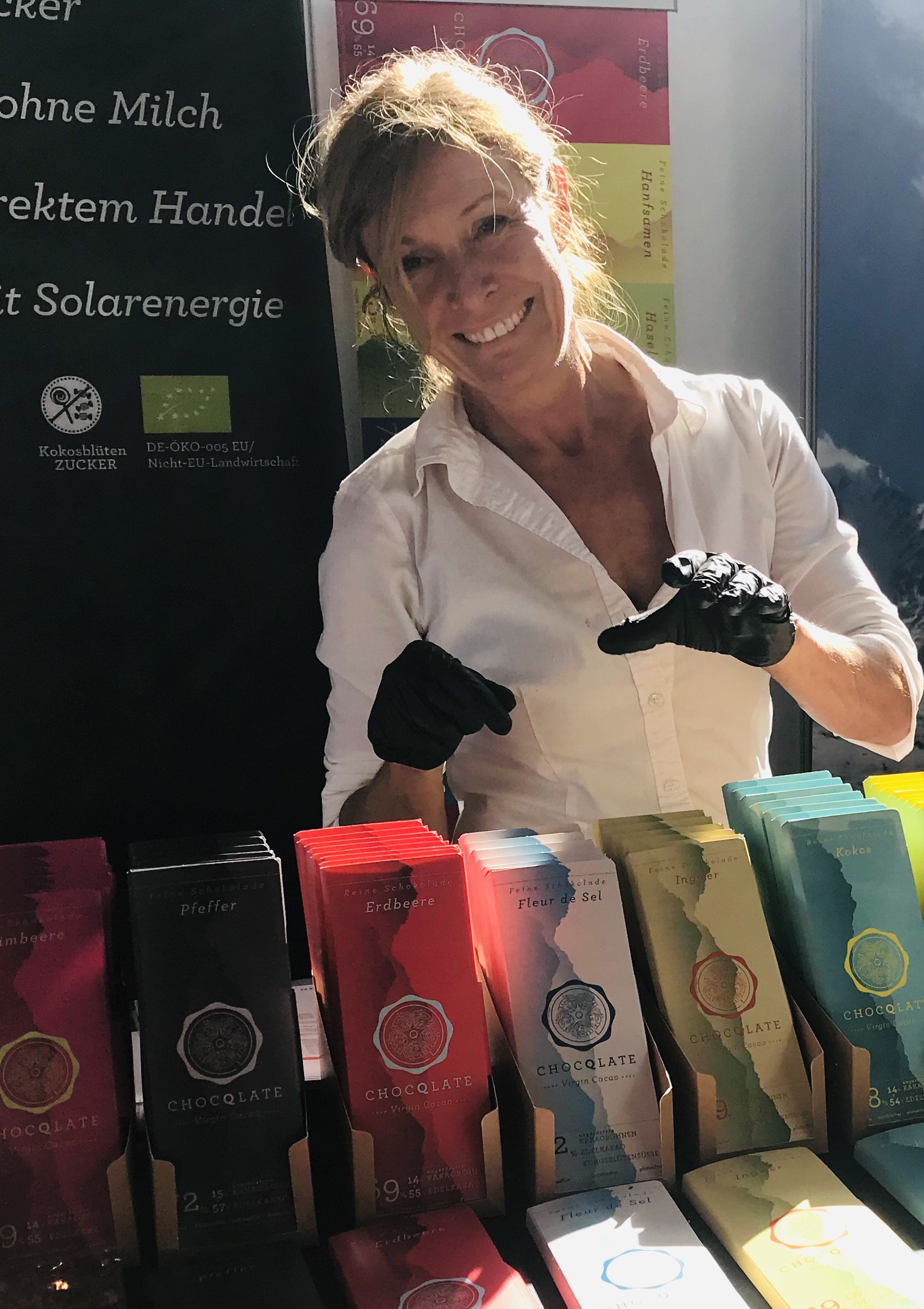 Porträt von Antonie: lachende Frau mit blondem, zurückgebundenem Haar und weißer Bluse im Sonnenlicht an einem Marktstand, vor ihr bunte CHOCQLATE-Tafeln in Kraftkarton-Displays