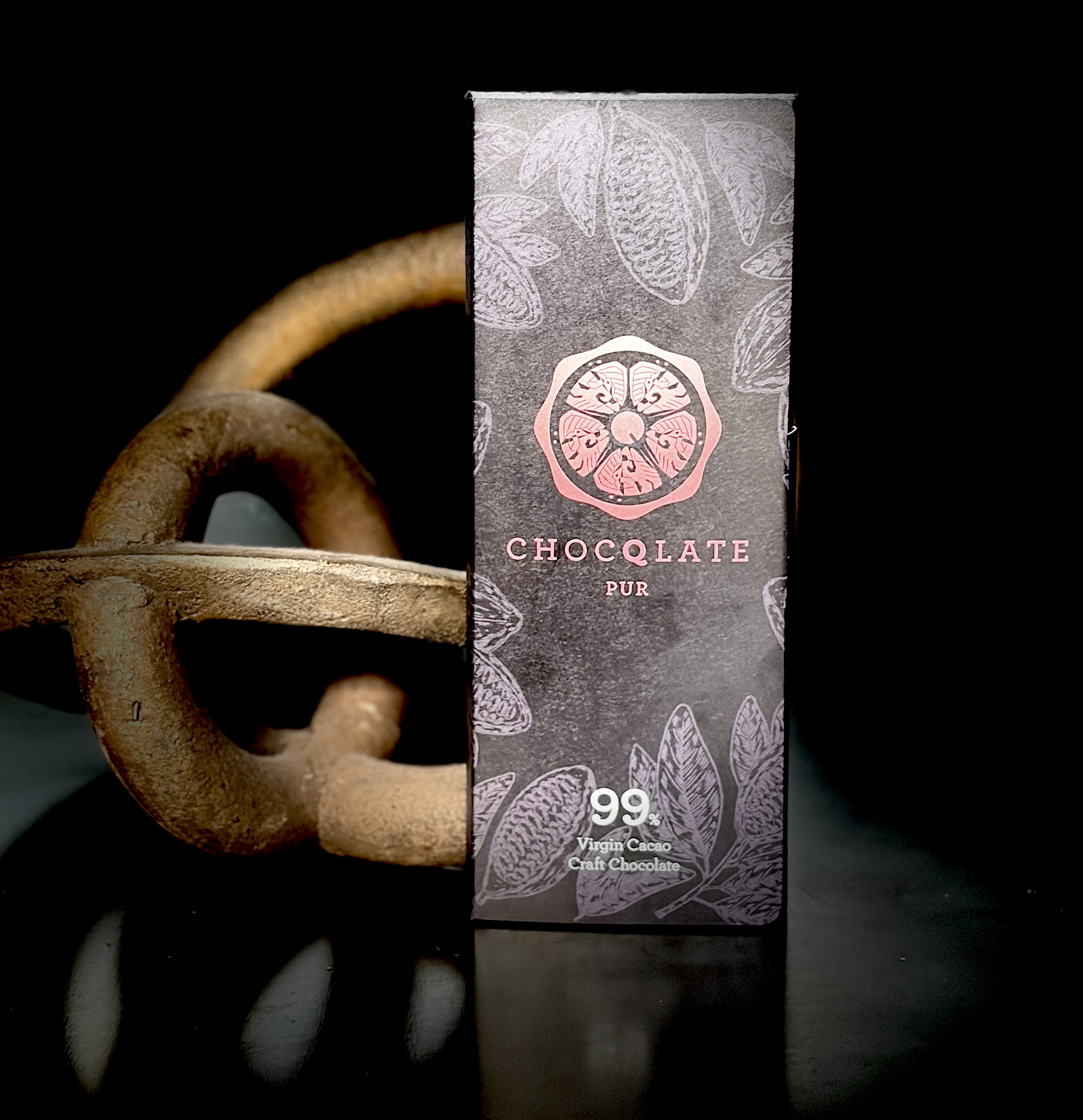 CHOCQLATE Pur 99 % Virgin Cacao Craft Chocolate vor einer rostigen Eisenskulptur auf dunklem Grund