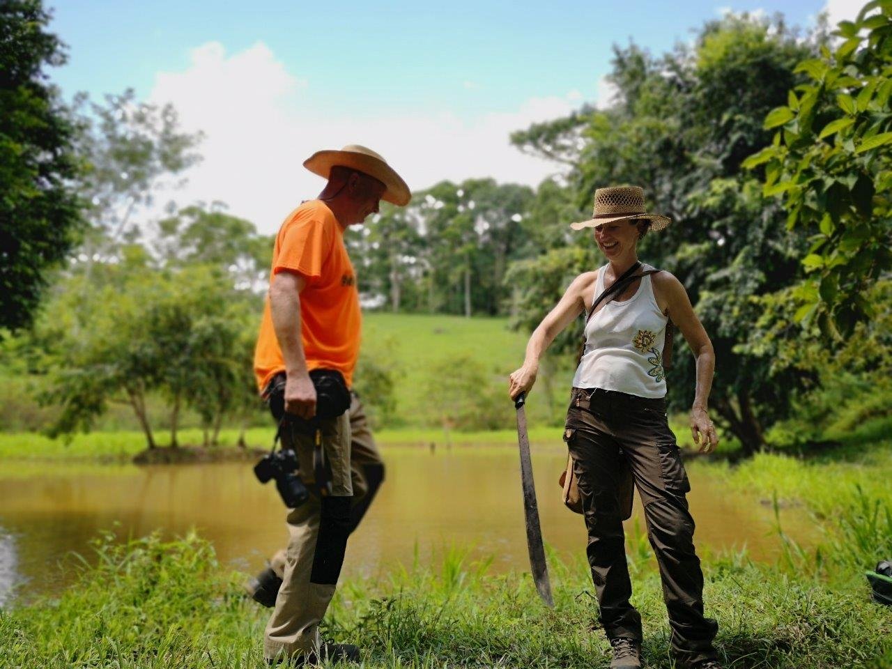 An einem Teich mit braunem Wasser stehen ein lachender Mann im orangefarbenen T-Shirt mit Hut und Kamera und eine lachende Frau mit Strohhut, die eine lange Machete auf den Boden stützt