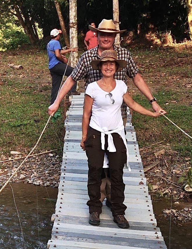 Eine lächelnde Frau mit Strohhut geht über eine schmale Hängebrücke aus Holzlatten, direkt hinter ihr hält ein Mann im karierten Hemd mit hellem Cowboyhut lachend die Seile