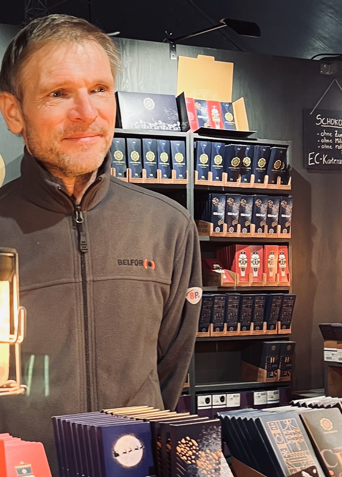 Porträt von Christian: Mann mit grau meliertem Haar und grauer Fleecejacke an einem CHOCQLATE-Marktstand, hinter ihm ein Holzregal voller Schokoladentafeln