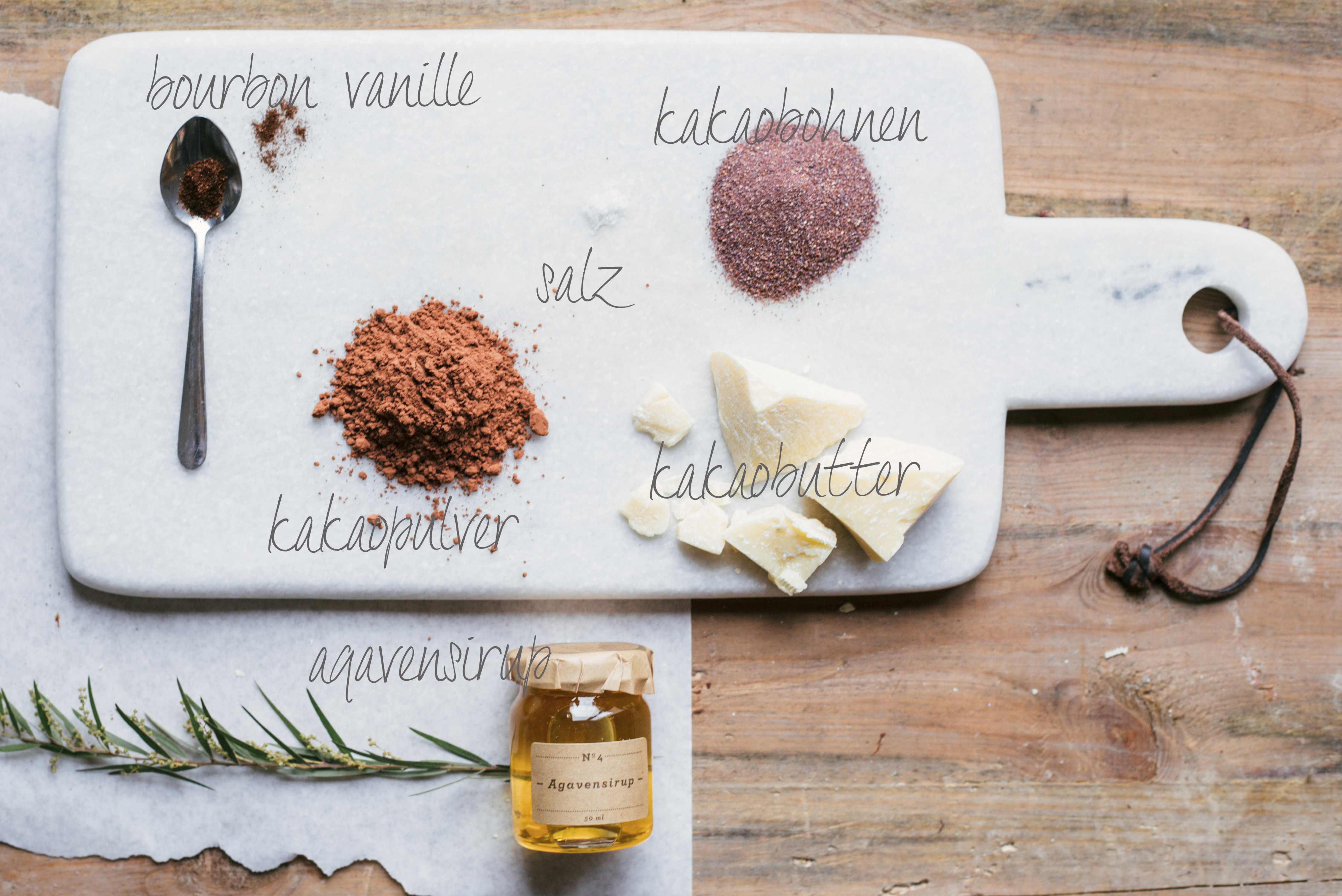 Zutaten für selbstgemachte Schokolade auf einem Marmorbrett: Kakaopulver, Kakaobohnen, Vanille, Salz, Kakaobutter und Agavensirup