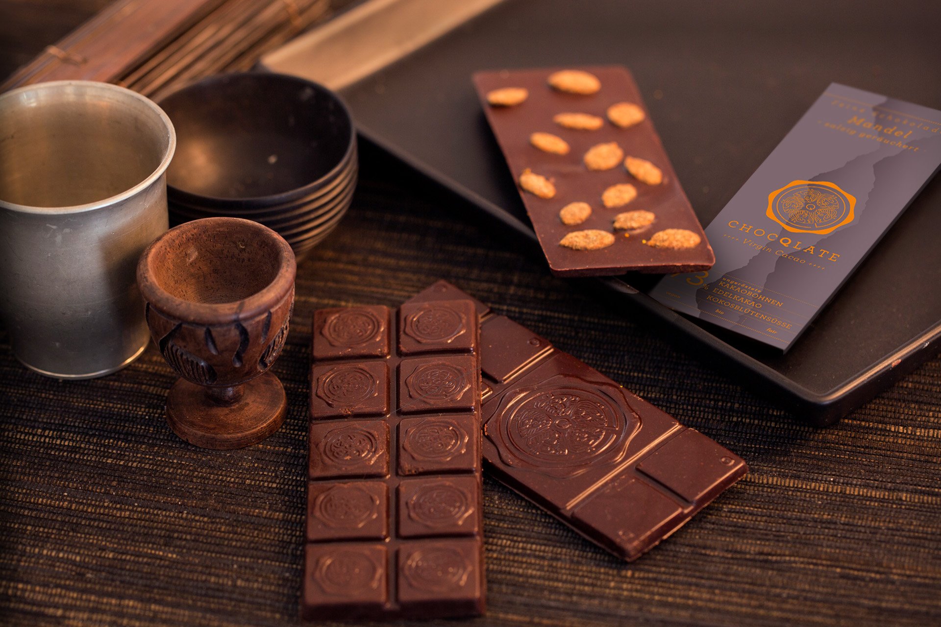 Frühe CHOCQLATE-Schokoladentafeln mit Siegelprägung, eine Tafel mit Mandeln und die historische Virgin-Cacao-Verpackung auf dunklem Holz, daneben Holzbecher, ein Metallbecher und dunkle Schalen