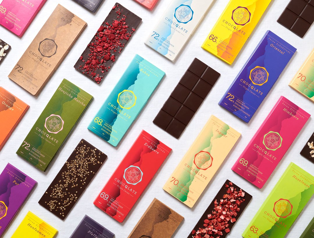 Farbenfrohes CHOCQLATE-Tafelsortiment auf hellem Leinen von oben: Verpackungen mit Farbverläufen und Siegel, Aufschrift „Feine Schokolade · Virgin Cacao“, dazwischen ausgepackte Tafeln, teils mit Himbeeren, Erdbeeren oder Hanfsamen bestreut