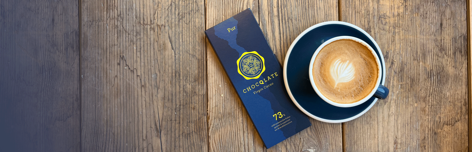 Eine Tafel CHOCQLATE „Pur“ 73 % in dunkelblauer Verpackung mit goldgelbem Siegel und dem Schriftzug „Virgin Cacao“ liegt neben einem Cappuccino auf verwittertem Holz
