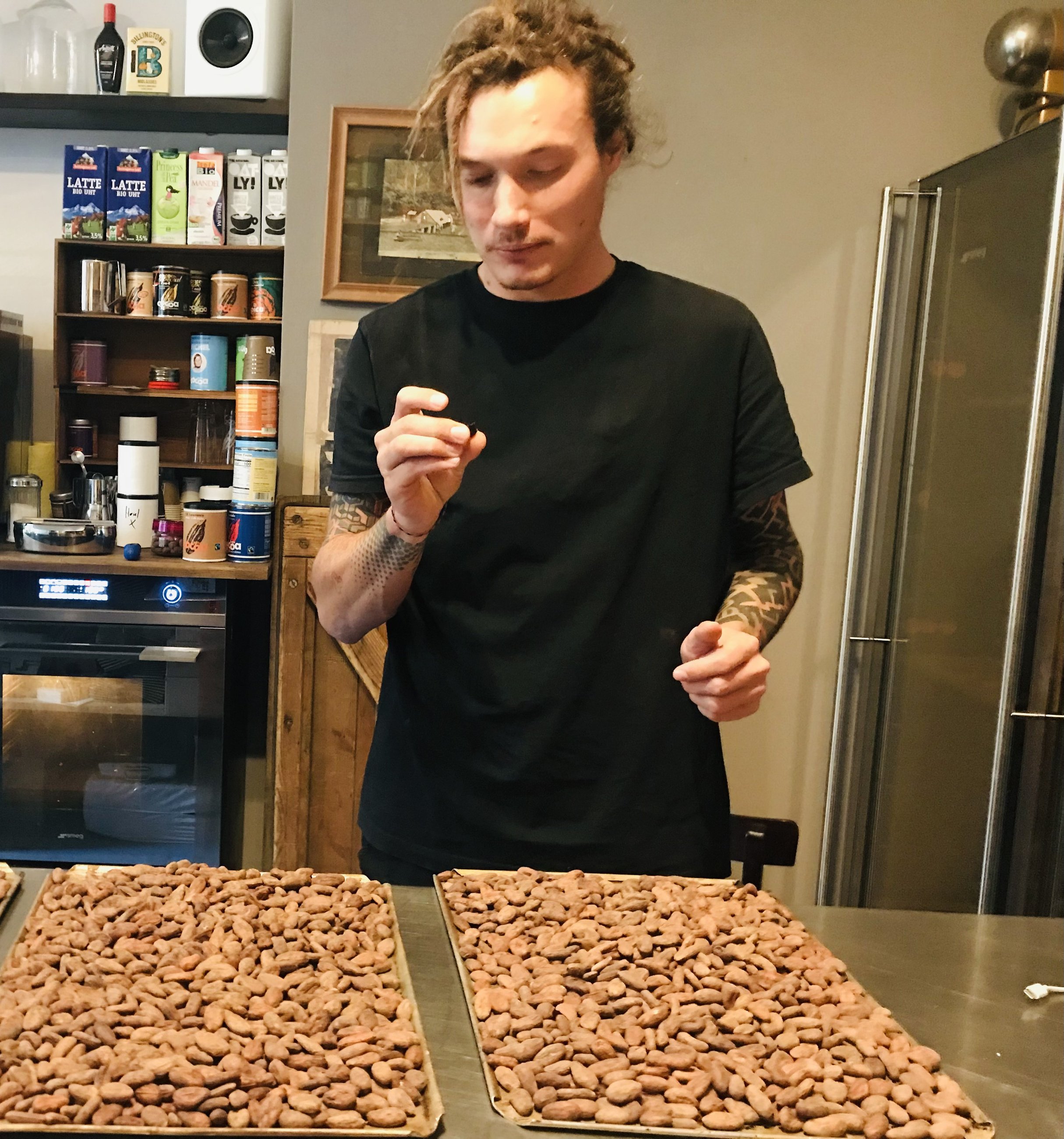 Porträt von Demian: junger Mann mit hochgebundenen Dreadlocks, schwarzem T-Shirt und tätowierten Unterarmen probiert konzentriert eine Kakaobohne, vor ihm zwei Bleche mit getrockneten Kakaobohnen